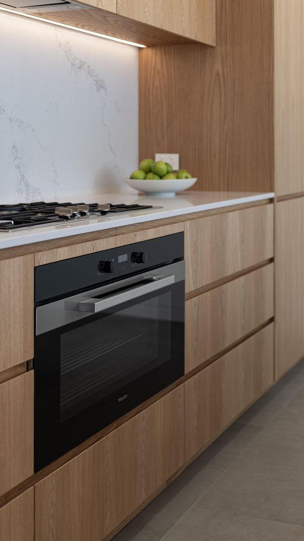 Close-up view of a contemporary kitchen featuring warm light-wood cabinetry, integrated drawers, a black built-in oven, gas cooktop, pale veined stone backsplash and benchtop, and subtle under-cabinet lighting. The composition focuses on the fitted kitchen joinery and appliances.