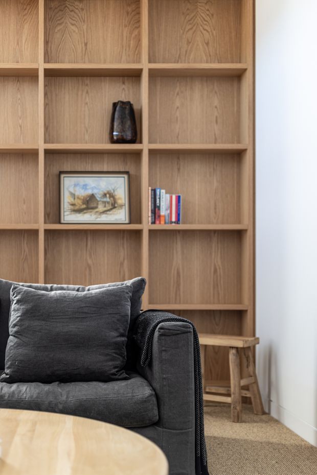 A softly lit living room corner featuring a dark charcoal upholstered armchair or sofa in the foreground, a full-height built-in timber bookcase with open square shelving, and a small wooden stool tucked beside it. The shelving displays a few books, a framed artwork, and a decorative vessel. A light timber round coffee table and neutral textured carpet complete the warm, restrained interior.