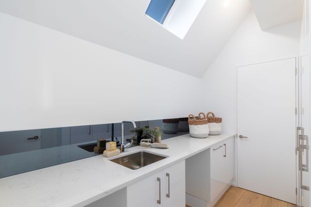 Bright, compact contemporary kitchen with white cabinetry and a white stone benchtop, a stainless-steel undermount sink, mirrored blue-grey splashback, pale timber flooring, and a sloped ceiling with skylight. Woven baskets and small plants add subtle natural texture.