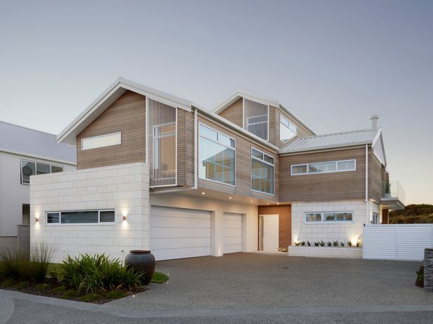 Contemporary multi-level residential home viewed from the street, featuring intersecting pitched gabled roof forms, extensive glazing, warm horizontal timber cladding, white masonry walls, integrated garages, and a broad paved driveway with landscaped planting.