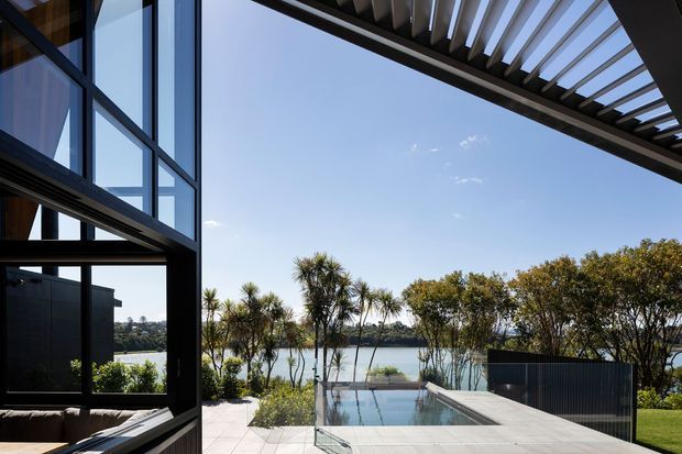 Contemporary waterfront residence viewed from an outdoor terrace, with expansive dark-framed glazing, a projecting pergola or louvred canopy, pale paved flooring, and a rectangular swimming pool overlooking a calm body of water. The image emphasises indoor-outdoor connection, strong geometric lines, and a bright landscaped setting.