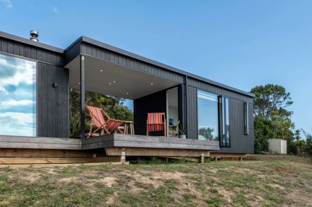 Exterior view of a compact modern single-storey house with dark vertical cladding, a low-pitched roof, extensive glazing, and a recessed covered outdoor deck containing chairs. The building is elevated slightly above a sparsely landscaped site.