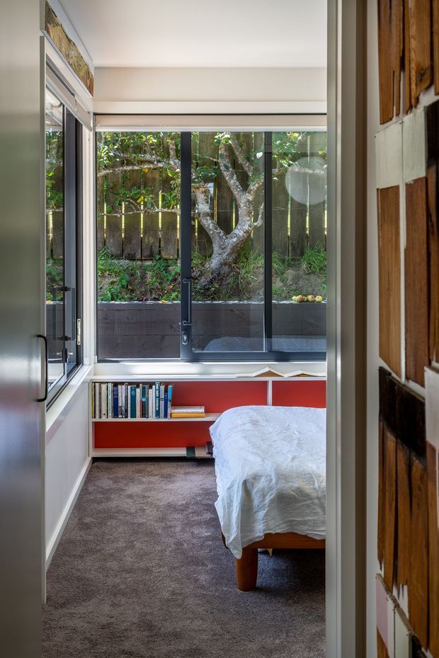 Compact bedroom viewed through an open doorway, with a partially visible bed, wall-to-wall grey carpet, white walls, and a low built-in red open bookshelf beneath large black-framed windows. The windows bring in abundant natural light and look onto a planted garden with a mature tree. A timber-panelled door or wall is visible at the right edge.
