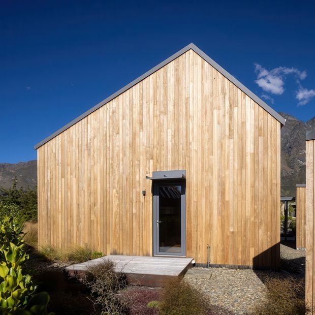 Exterior view of a simple gable-roofed building clad in vertical natural timber boards, with a dark framed entry door and a small concrete landing. The structure sits in a rugged mountainous landscape under a clear blue sky, with gravel and drought-tolerant landscaping in the foreground.