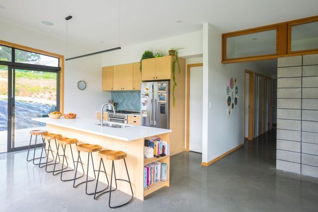 Bright open-plan kitchen with a long light-wood island and integrated seating for four, pale timber cabinetry, stainless-steel appliances, concrete-look flooring, and a white ceiling. A glazed exterior door/window brings in abundant daylight, while a hallway and textured block feature wall are visible beyond the kitchen.