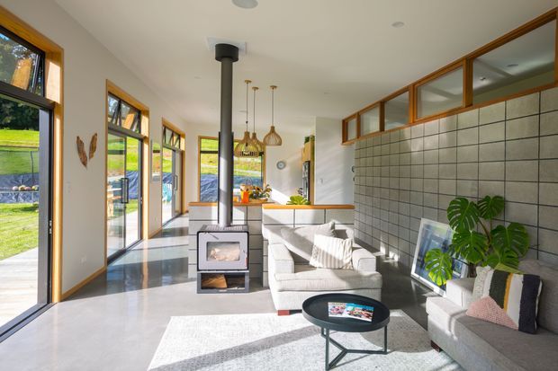 Bright contemporary open-plan living area with polished concrete flooring, a tiled feature wall, timber-framed glazing and clerestory windows. A compact wood-burning stove sits centrally between the lounge and kitchen, with pale upholstered seating, pendant lights and views to a green outdoor setting.