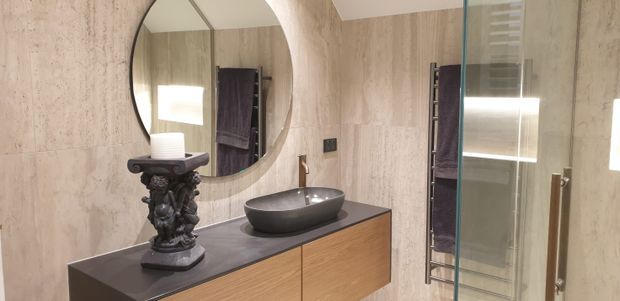 A refined contemporary bathroom featuring large beige stone-look wall tiles, a dark stone vanity benchtop, warm timber cabinetry, a round wall mirror, vessel basin, sculptural dark decorative lamp, and a glass-enclosed shower with brass-toned hardware.