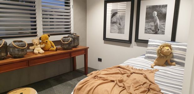 A cozy children's bedroom with a single bed dressed in grey-and-white striped bedding and a warm tan blanket. Plush toys sit on the bed and on a wooden console beneath horizontal window blinds, with framed black-and-white artwork on the wall and dark flooring.