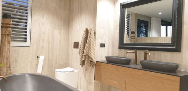 A warm, contemporary bathroom featuring a freestanding bathtub in the foreground, a wall-mounted timber vanity with two vessel basins, brushed metal tapware, a framed mirror, a toilet, and beige textured wall finishes. A window with horizontal louvres provides natural light.