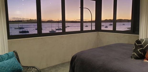 A bedroom with a large corner arrangement of dark-framed windows overlooking a calm waterfront with moored sailboats at dusk. The room contains a bed with dark bedding, a patterned accent cushion, and a woven lounge chair with a turquoise cushion. Neutral walls, carpet, and soft curtains create a quiet, relaxed atmosphere.