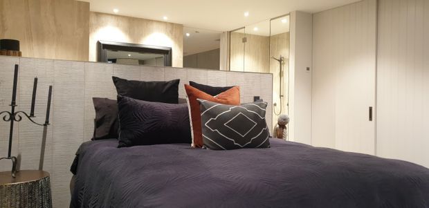 A contemporary bedroom viewed from the foot of the bed, featuring dark purple bedding and layered decorative cushions. Behind the bed is a textured light-grey upholstered headboard or partition, with warm timber wall panels, mirrored surfaces, recessed ceiling lights, and a partially visible glass-enclosed bathroom with shower. Minimal white cabinetry and doors complete the room.