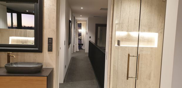A refined bathroom vanity area opens onto a long interior hallway. The bathroom features a vessel basin on a timber-toned vanity, brushed brass tapware, a large dark-framed mirror, and a tall illuminated stone-look cabinet or feature panel with brass handles. The hallway beyond has dark carpet, white walls, recessed ceiling lights, and dark balustrade-style joinery.