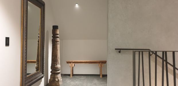 A quiet upper-level landing or hallway with a large ornate dark-framed mirror on the left, a carved timber decorative post, a simple wooden console table, grey carpet, textured light walls, recessed ceiling lighting, and a black metal stair balustrade descending at right.