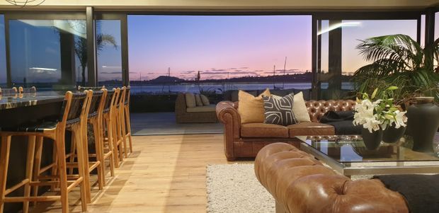 A spacious open-plan living and dining area with a kitchen island and timber bar stools on the left, a tufted brown leather sectional and sofa in the foreground, and a glass coffee table. Full-height glazing opens to a waterfront outlook at dusk, while large indoor plants and warm timber flooring add a relaxed, refined character.