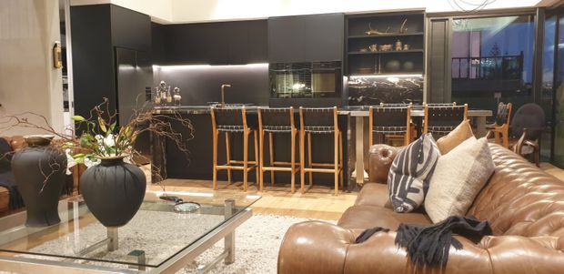 Open-plan contemporary kitchen and living area with a dark charcoal kitchen, central island and timber bar stools, a large tan leather sectional sofa, glass coffee table, and patterned rug. Built-in illuminated shelving and glazing add depth, while decorative plants and cushions soften the space.