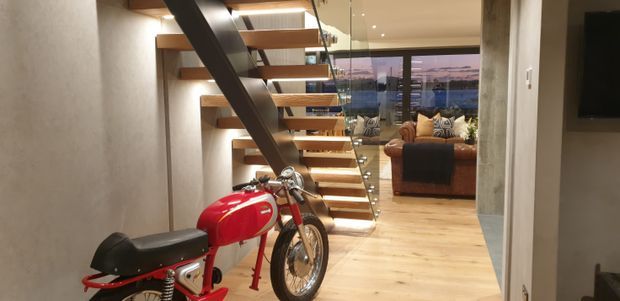 Interior view of a contemporary home featuring a red motorcycle displayed in the entry area, a floating timber staircase with a dark structural beam, warm timber flooring, and a living room beyond with expansive glazing and a sofa.