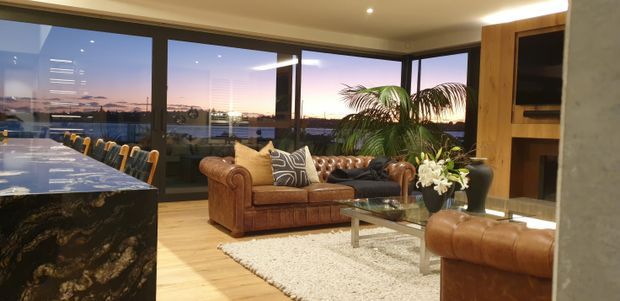A spacious, warmly lit open-plan living area with floor-to-ceiling corner glazing overlooking a sunset landscape. The room features a large dark patterned stone kitchen island, light timber flooring, a deep brown Chesterfield-style leather sofa and ottoman, a glass coffee table, indoor palms, and built-in timber cabinetry with a television and fireplace area.