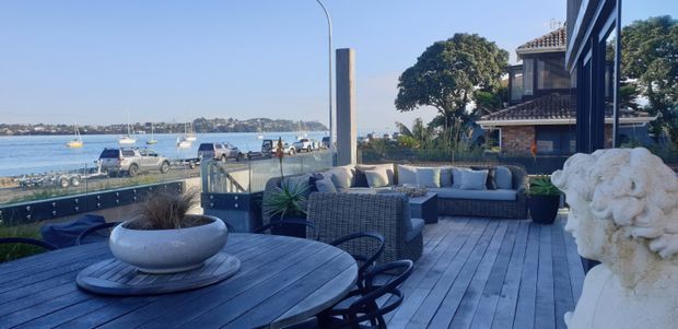 A spacious waterfront outdoor deck or terrace furnished with a round timber dining table, dark woven chairs, and a large outdoor sectional sofa. The deck overlooks a marina or bay with boats and a tree-lined shoreline. A substantial sculptural stone feature sits at the right edge, while a dark-clad building and upper-level tiled roof are visible behind the terrace.