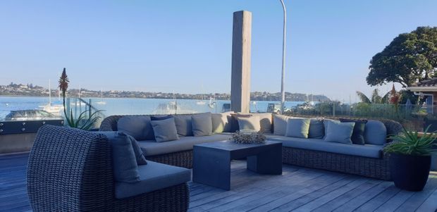 A stylish waterfront outdoor lounge terrace with woven modular sofas and armchairs arranged around a low square coffee table. The space is finished with grey timber decking, pale cushions, large potted plants, a sculptural timber post and slender curved light, with expansive water and distant shoreline views beyond the glass balustrade.