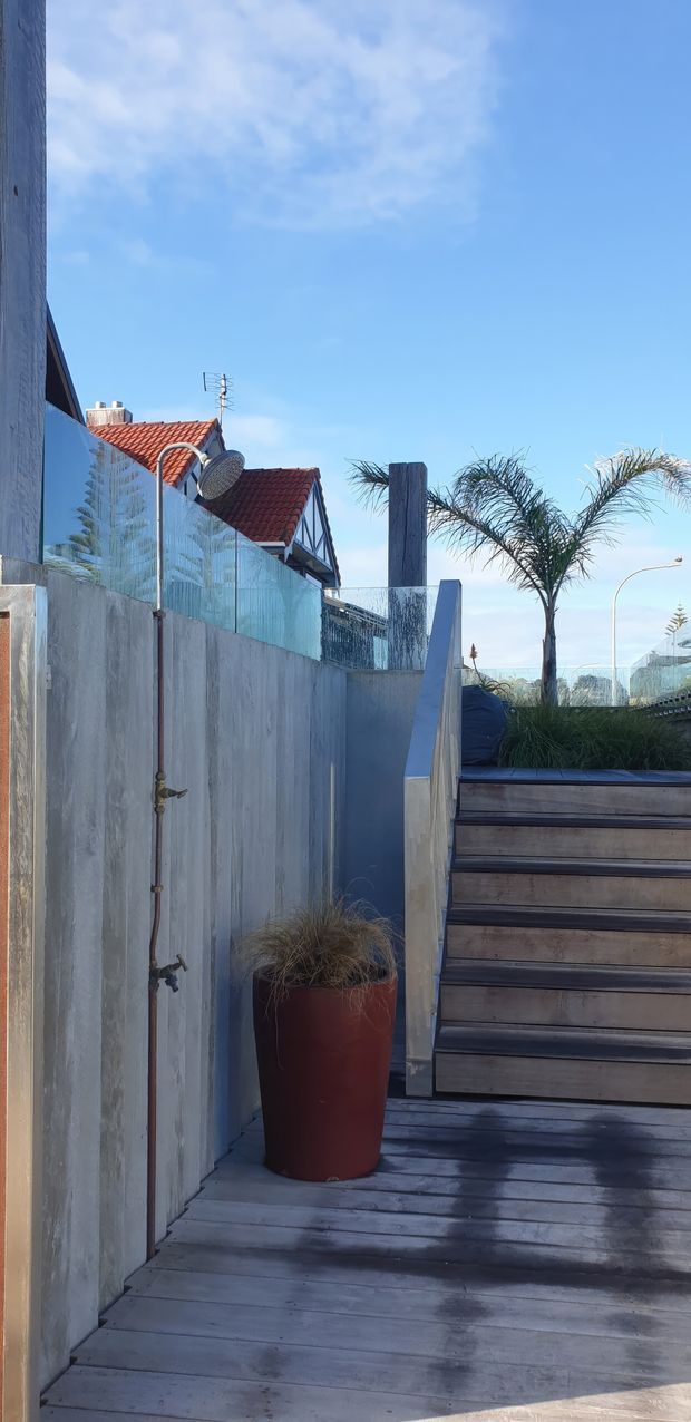 Vertical photograph of a compact outdoor deck and stairway beside a rendered concrete wall. The deck and steps are finished in weathered grey timber, with a tall terracotta planter, ornamental grasses, palm trees, and an exposed outdoor shower. A glass or transparent privacy screen borders the upper deck, while a neighbouring red tiled roof and blue sky are visible beyond.