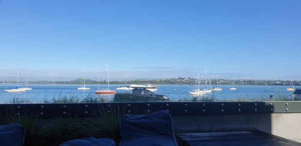 A waterfront outdoor terrace or balcony overlooking a calm bay with moored sailboats and a distant shoreline. The foreground contains dark built-in seating or cushions, ornamental grasses, and a low dark metal-clad privacy wall with visible fasteners. The scene is bright, open, and coastal, with blue water and sky dominating the view.