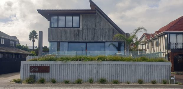 Street-facing exterior view of a contemporary two-storey house with a steep asymmetrical gable roof, vertically clad weatherboard facade, dark-framed glazing, and a landscaped raised front garden behind a low concrete or rendered boundary wall. Neighbouring residential homes are visible on either side.