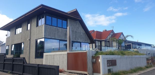 Street-facing view of a contemporary two-storey home with a steep gabled roof, weathered vertical timber cladding, broad dark-framed glazing, concrete boundary walls, and a warm timber gate. Adjacent homes with pitched roofs are visible in the background, with coastal-looking sky and planting around the property.