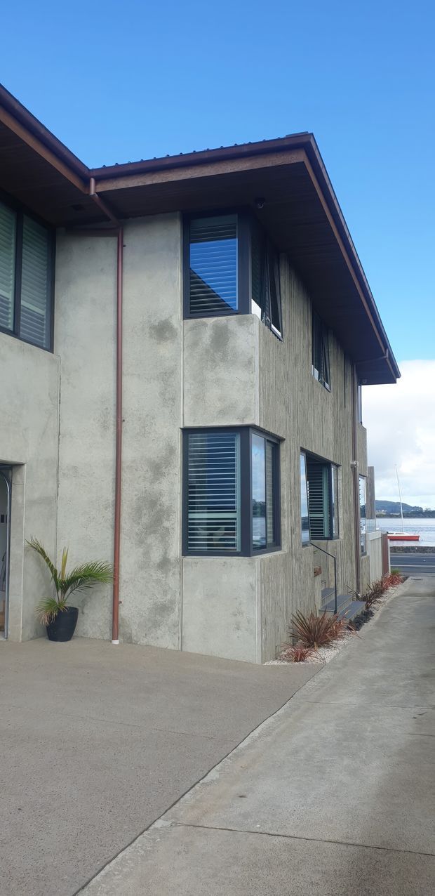 Exterior view of a multi-level coastal building with a low-pitched roof, deep timber-lined eaves, textured concrete and vertically ribbed wall surfaces, and dark-framed louvred windows. A paved access area and landscaped planting border the building, with a marina or waterfront visible beyond.