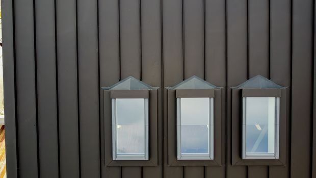 Close-up exterior view of a dark vertically clad wall with three evenly spaced, small projecting lantern-like windows or glazed wall fixtures. The narrow rectangular units have peaked caps and dark frames, creating a repetitive geometric facade detail.