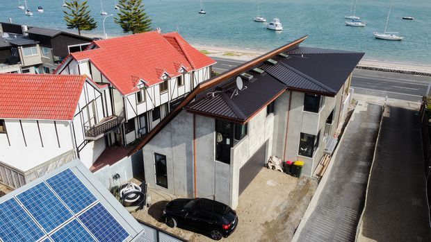 Aerial view of a compact coastal residential development beside a waterfront road, showing a contemporary multi-level building with dark metal pitched roofing, concrete walls, large windows, rooftop solar panels, and adjacent Tudor-style homes with red tiled roofs. Boats are visible on the bay beyond.