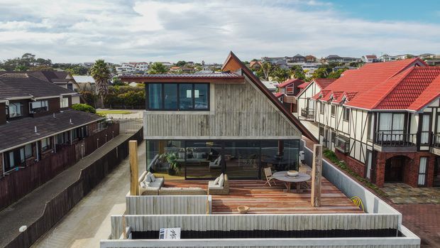 Elevated exterior view of a contemporary multi-level home in a dense suburban setting. The house features a steep gabled roof, weathered vertical timber cladding, extensive dark-framed glazing, and a large timber rooftop deck with outdoor seating. Surrounding homes and red-roofed neighbouring buildings are visible.
