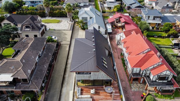 Aerial view of a suburban residential neighbourhood showing several detached houses, varied pitched and gabled roofs, landscaped yards, driveways, and a prominent contemporary home with a dark metal roof and timber outdoor terrace.
