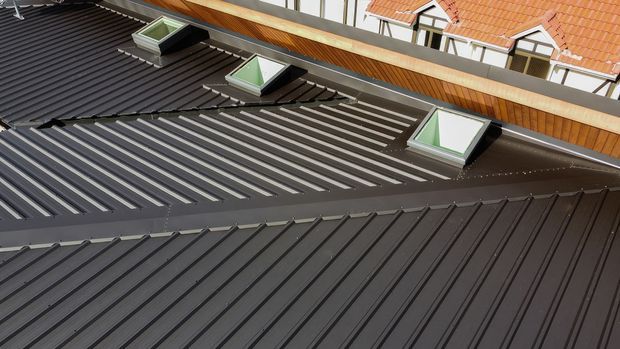 Close-up exterior view of overlapping dark metal roofing, featuring multiple raised seams and several angled skylights. A terracotta-tiled roof and light-coloured building elements are visible in the background.