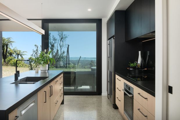 Contemporary galley kitchen with a large central island, pale timber cabinetry, dark charcoal benchtops and overhead joinery, integrated stainless-steel appliances, and black-framed full-height glazing. The glazing provides views toward a coastal landscape and adds abundant natural light. A polished concrete or terrazzo-like floor and minimalist fittings complete the space.