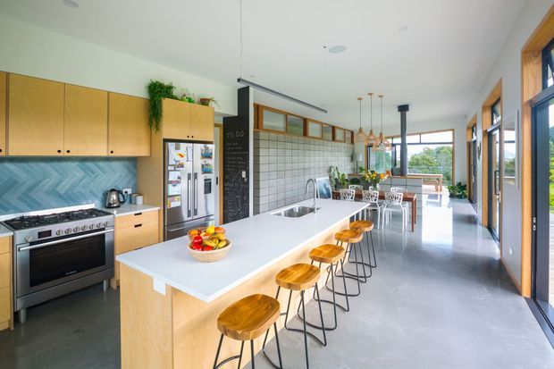 Bright, spacious open-plan kitchen and dining area with a long pale timber island, white benchtop, integrated sink, and multiple timber stools. The kitchen includes full-height timber cabinetry, a stainless-steel range cooker and refrigerator, and a blue-grey herringbone tile splashback. A tiled feature wall, pendant lights, dining table, and large timber-framed windows complete the airy contemporary space.