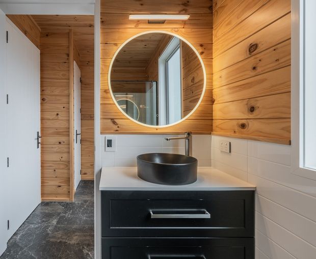 Compact contemporary bathroom vanity area with a black vessel basin on a white benchtop, dark drawer cabinetry, white horizontal wall tiles, and warm timber wall and ceiling panelling. A large round mirror reflects the timber-lined interior and window, while a white door and dark stone-look floor are visible to the left.