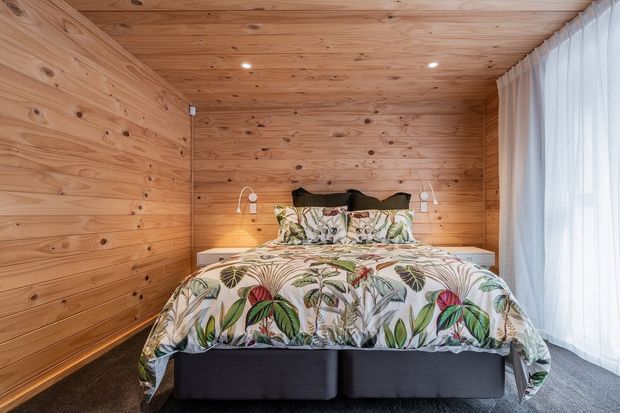A compact bedroom fully lined in warm natural timber, with a double bed, dark upholstered base, botanical-patterned bedding, recessed ceiling lights, wall-mounted reading lamps, and sheer curtains covering a large window.