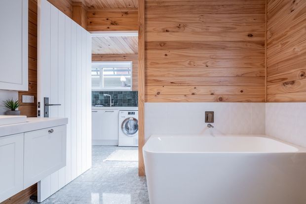 Bright contemporary bathroom with a freestanding white bathtub, white wall-hung vanity, and extensive warm timber wall and ceiling panelling. A laundry with white cabinetry and a front-loading washing machine is visible through the open doorway. The bathroom has a polished grey concrete-look floor, white tiled lower walls, and simple black fixtures.