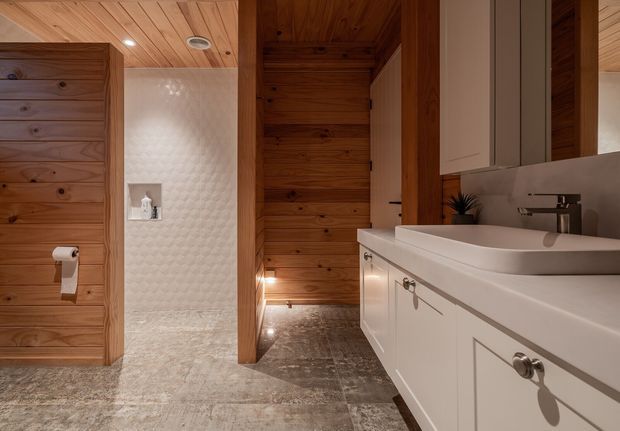 Warm contemporary bathroom featuring extensive timber wall and ceiling panelling, a textured light tiled shower enclosure, stone-look floor tiles, and a long white vanity with integrated basin, framed cabinetry, chrome tapware, and mirrored storage.