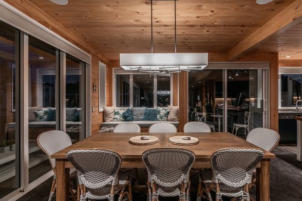 A warm, contemporary dining area with a large rectangular timber dining table surrounded by woven cane dining chairs. The room features extensive dark-framed glazing and sliding doors, a timber-lined ceiling with exposed beams, a large rectangular pendant light, and views into adjacent living and kitchen spaces.