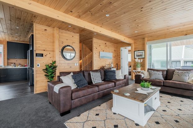 Warm open-plan living room with extensive timber wall and ceiling lining, exposed timber beam, dark kitchen cabinetry, a brown sectional sofa, patterned rug, and large windows. A timber-lined hallway and stair area are visible behind the seating.