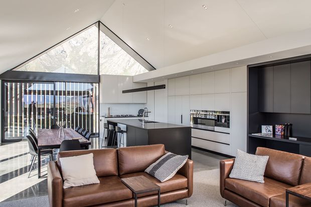 Open-plan contemporary living, dining, and kitchen interior with a dramatic triangular glazed gable and extensive vertical screening at the rear. The room features white full-height kitchen cabinetry, a dark island and dark feature joinery wall, integrated appliances, a timber dining table, and brown leather sofas with neutral cushions. Recessed ceiling lighting and clean-lined built-in elements create a refined, spacious interior.