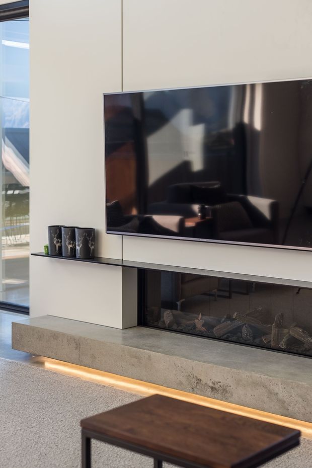 Contemporary living room media wall featuring a large wall-mounted television above a linear glass-fronted fireplace. The installation combines pale built-in wall panels, a dark slim shelf, a substantial concrete hearth, and warm recessed lighting. Floor-to-ceiling glazing is visible at the side, with a dark timber coffee table in the foreground.
