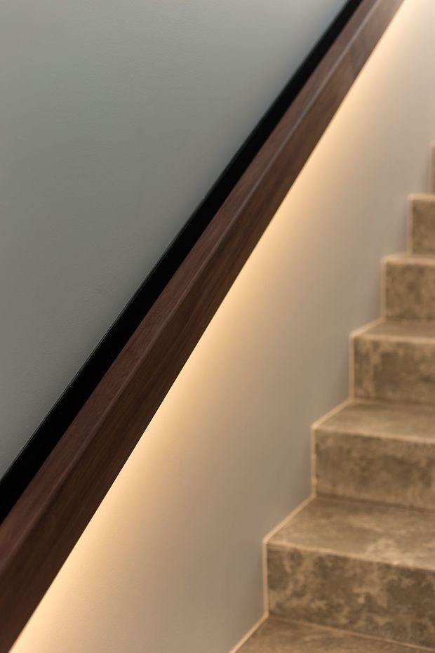 Close-up architectural detail of a staircase with warm integrated lighting beneath a dark timber handrail. The stair treads and risers are finished in mottled beige-grey stone or tile, while the adjacent wall is painted a warm light neutral. The composition highlights the handrail, concealed LED glow, and stair geometry.