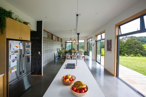 Bright open-plan kitchen dominated by a long white island with integrated double sink and seating for several people. The room has polished dark concrete flooring, warm timber-framed windows and doors, white walls, extensive glazing opening to a lawn, timber cabinetry, a stainless-steel refrigerator, and pendant lighting. A tiled feature wall and chalkboard panel add visual interest. The space feels spacious, airy, and family-oriented.