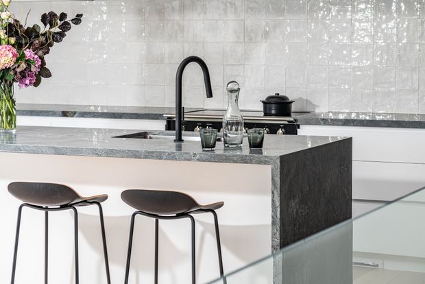 Bright contemporary kitchen featuring a large central island with a dark stone waterfall end, pale cabinetry, a black gooseneck tap, and a glossy white tiled splashback. Two dark stools provide island seating, while glassware, a carafe, cookware, and a floral arrangement add styling details.