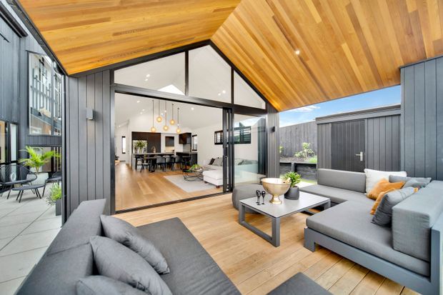 A contemporary covered outdoor lounge and entertaining area opening directly into a bright open-plan living and dining space. The room features extensive black-framed glazing, charcoal vertical wall cladding, warm timber ceilings and decking, grey upholstered sofas, and a dark coffee table. A kitchen and dining area are visible beyond the open glass doors, while a private courtyard and utility enclosure sit to the right.