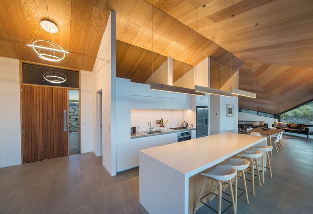 Open-plan contemporary kitchen and living area with a large white island, integrated cabinetry, tiled flooring, and timber-lined sloping ceilings. A vertical timber entry door and feature stone wall are visible, while linear pendant lights hang above the island. Glazing at the far end overlooks a green landscape.
