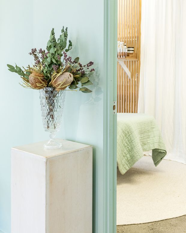 A softly styled pastel interior showing a mint-green painted wall and matching door frame, with a whitewashed pedestal topped by a glass vase of dried botanical foliage. Through the doorway is a calm bedroom with a pale green quilt, cream carpet, white curtains, and a timber slatted feature wall with built-in shelving.