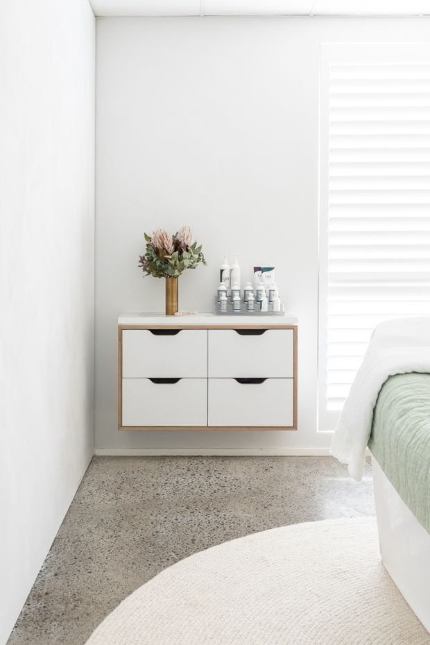 Bright, minimalist bedroom corner with a wall-mounted white and timber-toned drawer cabinet, toiletries and dried flowers on top, white walls, a large white shuttered window, pale green bedding, and a curved cream rug over a speckled concrete or terrazzo-style floor.